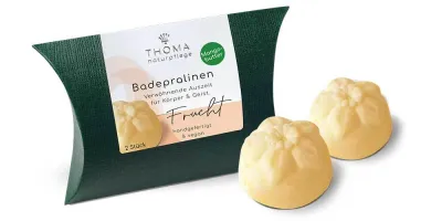 Zwei gelbliche Badepralinen vor dunkelgrüner Verpackung mit weißer Aufschrift Badepralinen Frucht, handgefertigt und vegan mit Mangobutter von der Naturseifen Manufaktur Uckermark.