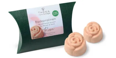 Zwei rosafarbene Badepralinen in Rosenform vor dunkelgrüner Verpackung mit weißer Aufschrift Badepralinen Rose, handgefertigt und vegan mit Sheabutter von der Naturseifen Manufaktur Uckermark.