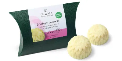Zwei cremefarbene Badepralinen vor dunkelgrüner Verpackung mit weißer Aufschrift Badepralinen Vanille, handgefertigt und vegan mit Sheabutter von der Naturseifen Manufaktur Uckermark.