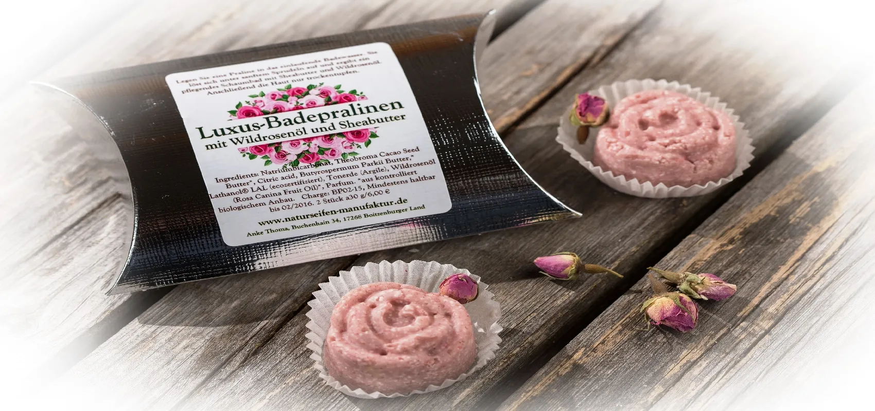 Luxus-Badepralinen mit Wildrosenöl und Sheabutter in einer schwarzen Verpackung, dekoriert mit getrockneten Rosenknospen, auf einem rustikalen Holztisch.
